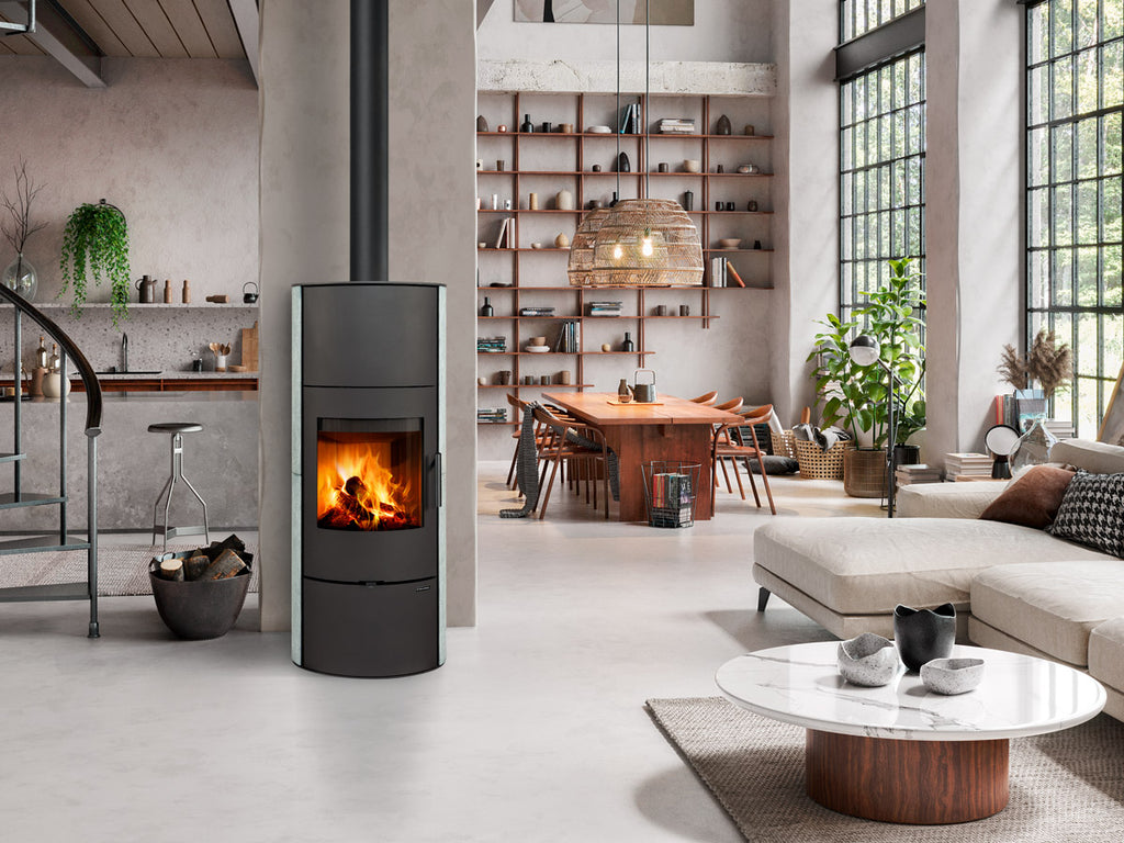 Sobă pe lemne modernă de tip turn cu uşă din fontă și sticlă curbată, integrată într-un living spațios tip loft cu decor industrial și zonă de dining.