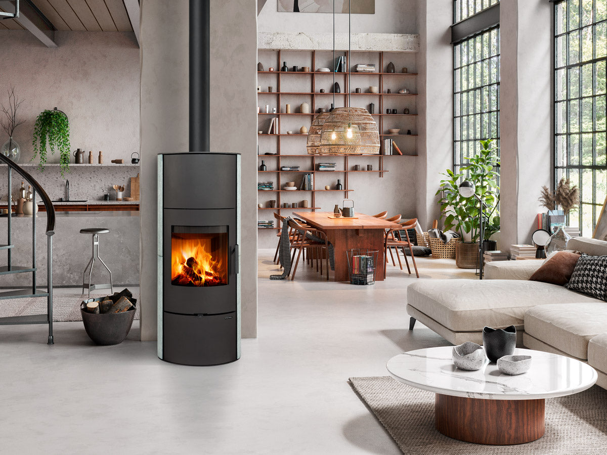 Sobă pe lemne modernă de tip turn cu uşă din fontă și sticlă curbată, integrată într-un living spațios tip loft cu decor industrial și zonă de dining.