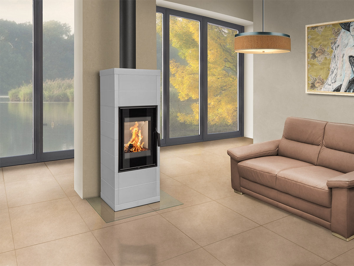 Living modern cu soba Fantasy 2 gri minimalista si foc aprins, canapea din piele maro, ferestre mari spre un lac si decor elegant in tonuri calde.