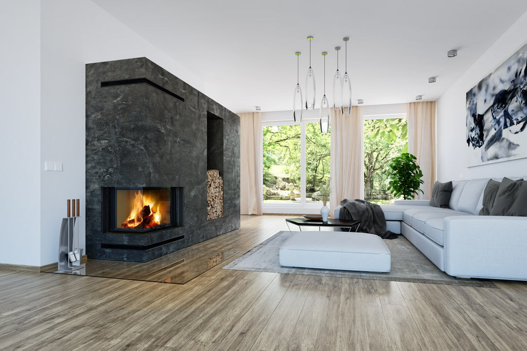 Living modern cu semineu pe lemne Romotop, colt stanga, integrat intr-un perete elegant finisat cu piatra neagra. Flacarile sunt vizibile prin sticla panoramica, oferind un efect spectaculos. Zona de relaxare include o canapea alba spatioasa, o masuta minimalista si un perete vitrat cu vedere catre gradina. Depozit pentru lemne integrat lateral, completand designul sofisticat al incaperii.