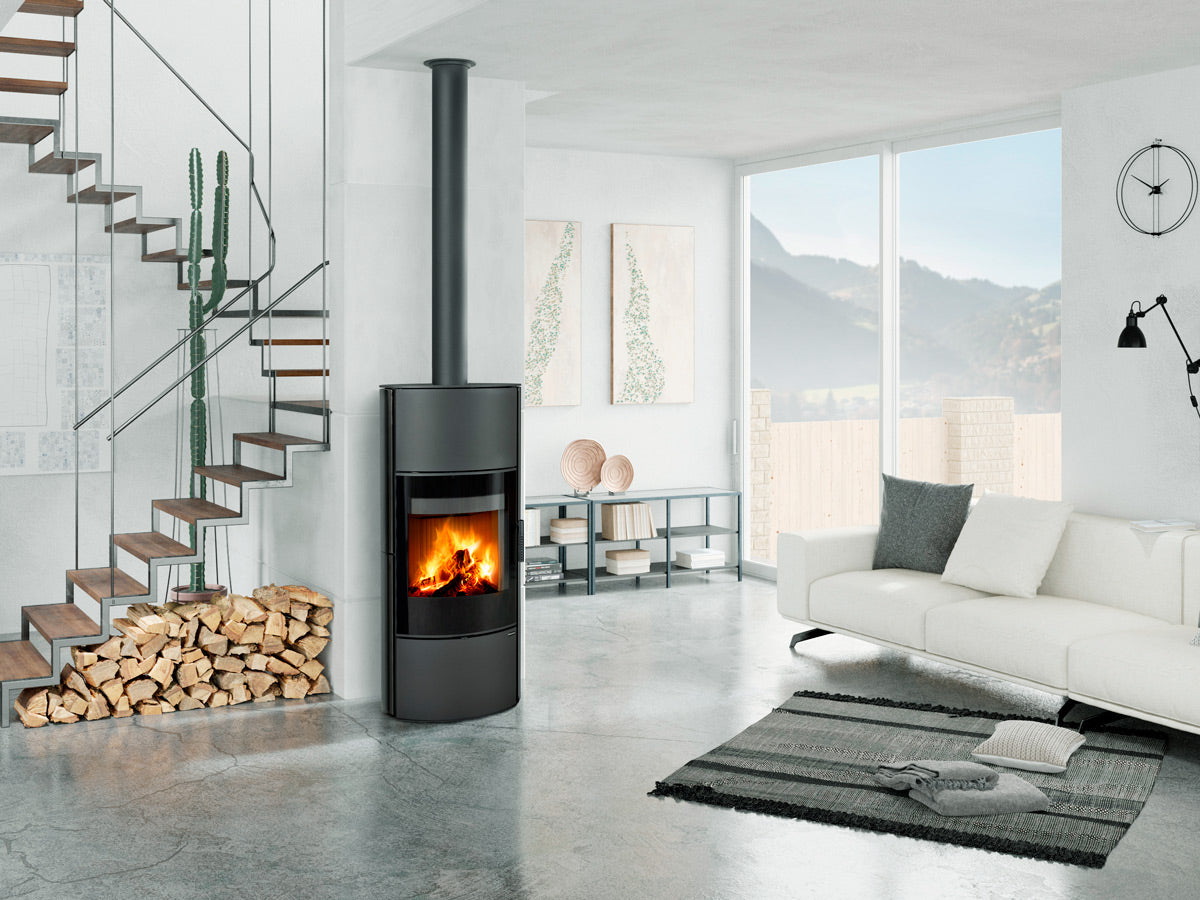 Soba ceramica Ovalis G H , cilindrica moderna cu geam panoramic si foc vizibil, instalata in living contemporan luminos, cu cos de fum vertical si depozitare lemne decorativa.