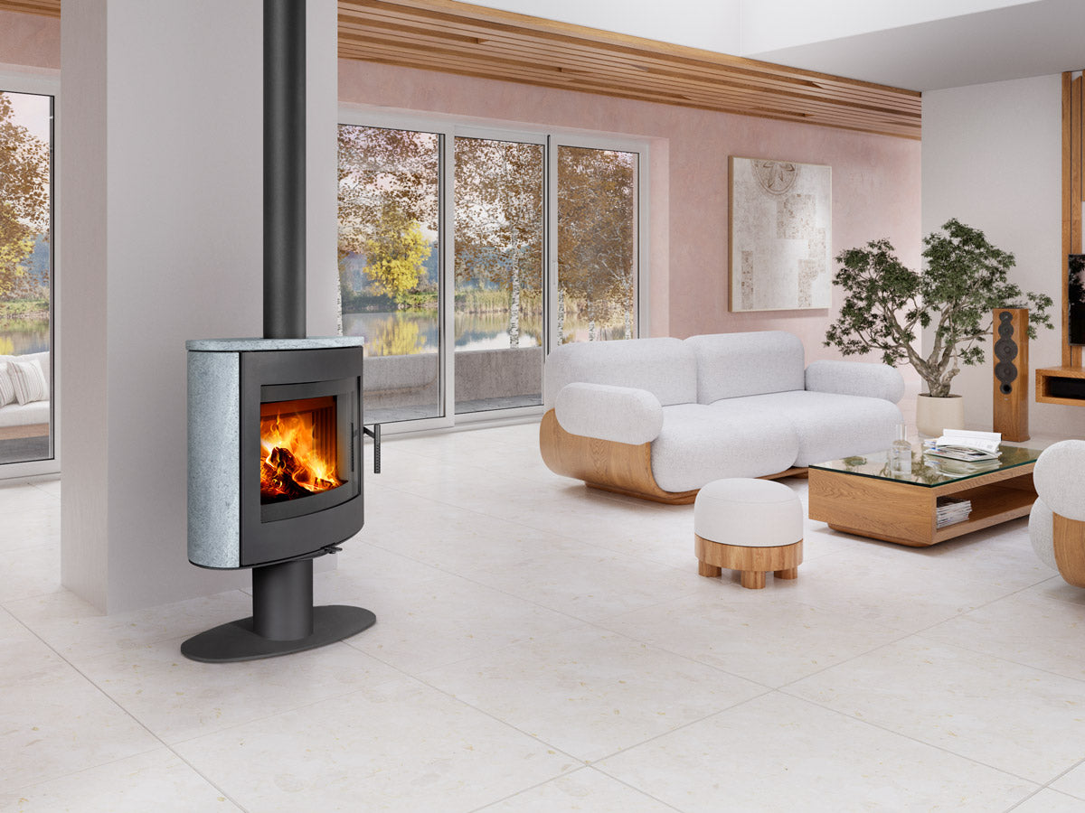 Soba lemne STROMBOLI T 20, forma rotunda, placata cu piatra gri si metal negru, cu usa de sticla si foc vizibil. Amplasata intr-un living modern, luminos.