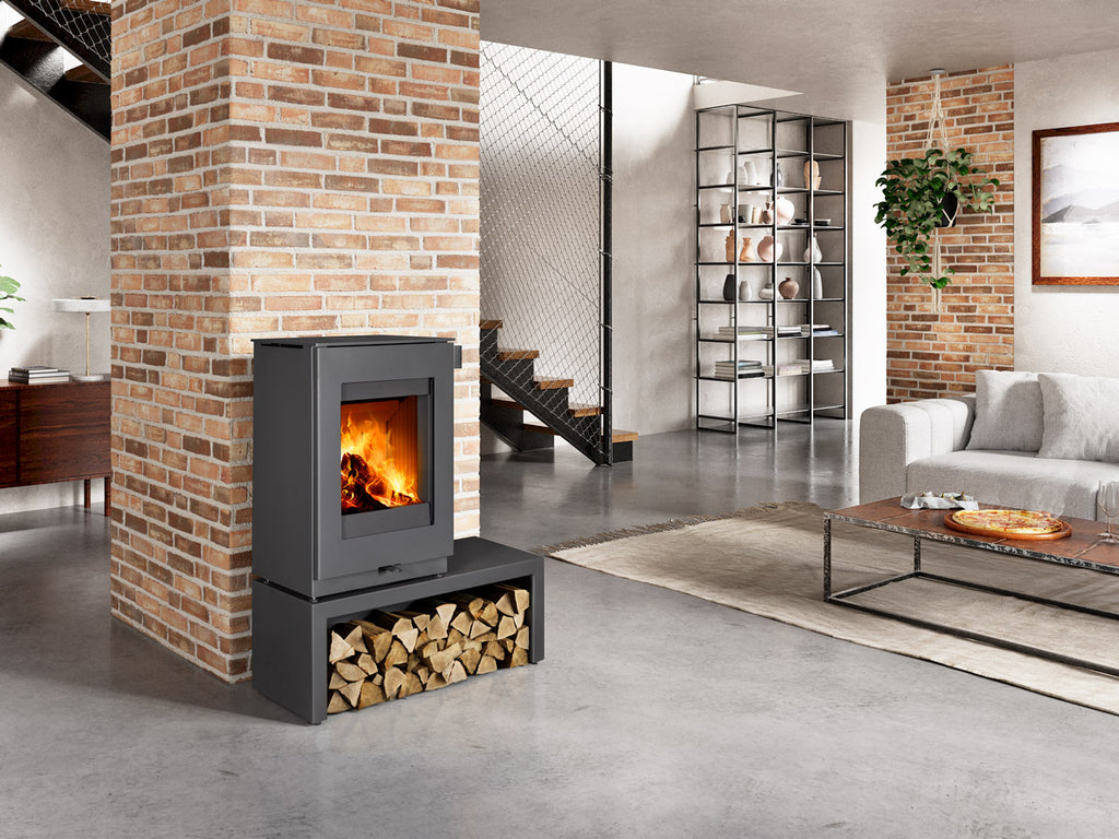 Soba lemne TEON P, metal antracit, cu geam frontal si foc vizibil. Include suport lemne. Amplasata intr-un living modern, stil industrial.